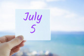 5. července. Ruka drží samolepku s textem 5. července na rozmazaném pozadí moře a oblohy. Kopírovat prostor pro text. Měsíc v koncepci kalendáře