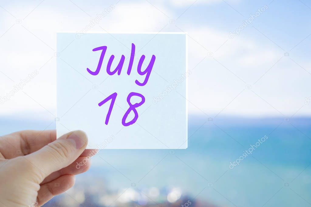 July 18th. Hand holding sticker with text July 18 on the blurred background of the sea and sky. Copy space for text. Month in calendar concept