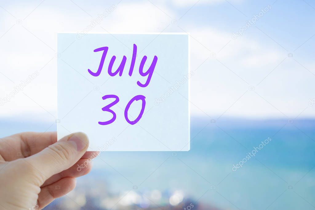 July 30th. Hand holding sticker with text July 30 on the blurred background of the sea and sky. Copy space for text. Month in calendar concept