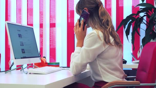 Mujer se sienta en el escritorio blanco con las conversaciones de ordenador en el teléfono toma notas en el cuaderno — Vídeo de stock