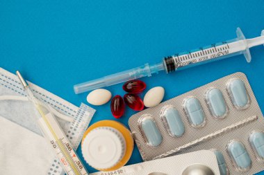 Tıbbi muayene ve tedavi konsepti. Kırmızı ve beyaz haplar, termometre, sarı serum, maske, mavi arka planda tıbbi şırınga fotokopi alanı. Tıbbi düzlük