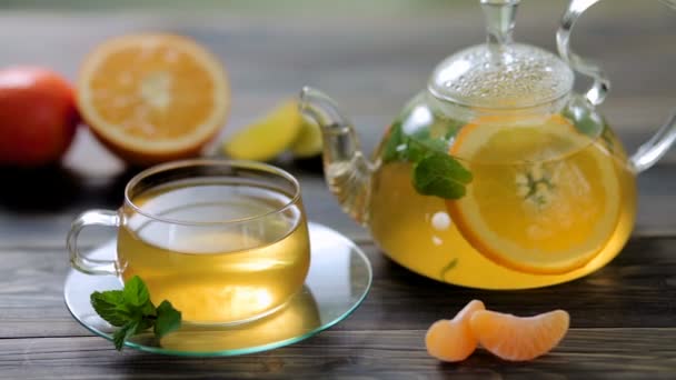 Teiera e tazza di vetro con tè verde, arancia, mandarino, lime e menta sul tavolo di legno marrone — Video Stock