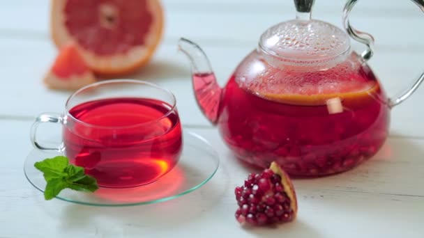 Théière en verre et tasse de thé rouge avec raisin, grenade, menthe sur une table en bois blanc — Video