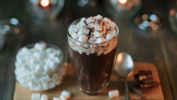 Gyönyörű kompozíció - forró csokoládé lekvárral és csokoládé darabok egy átlátszó pohár. Az üveg egy faállványon áll. A gyertyák mögött égnek. — Stock videók