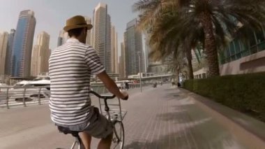 Genç bir adam bir şapka, çizgili t-shirt ve şort Promenade gökdelenler ve Dubai Marina, Birleşik Arap Emirlikleri yatlar arasında onun bisiklet sürme