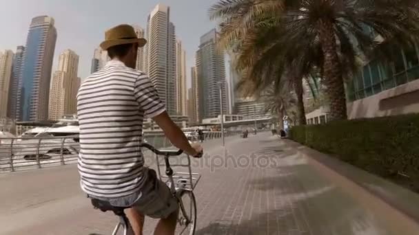Молодой человек в шляпе, футболке в полоску и шортах, катающийся на велосипеде по набережной среди небоскребов и яхт в Дубай Марина, ОАЭ — стоковое видео