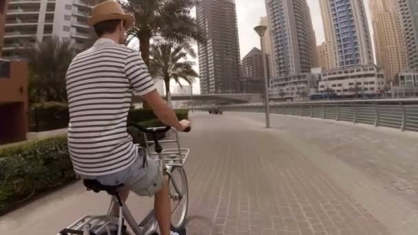 Молодой человек в шляпе, футболке в полоску и шортах, катающийся на велосипеде по набережной среди небоскребов и яхт в Дубай Марина, ОАЭ — стоковое видео