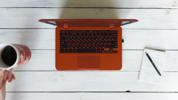 Hombre escribiendo texto en un portátil rojo que se encuentra en una mesa de madera blanca. A la izquierda está dando vueltas con un café negro, a la derecha hay un cuaderno — Vídeo de stock