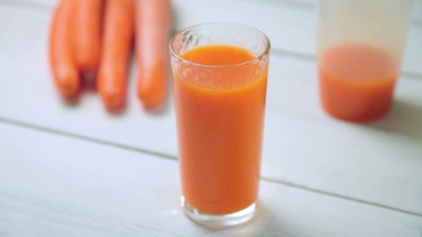 Склянка свіжого морквяного соку обертається навколо його осі на білому дерев'яному столі — стокове відео