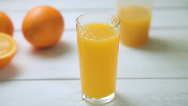 Ένα ποτήρι φρέσκο χυμό πορτοκάλι που περιστρέφεται γύρω από τον άξονά του σε λευκό ξύλινο τραπέζι — Αρχείο Βίντεο