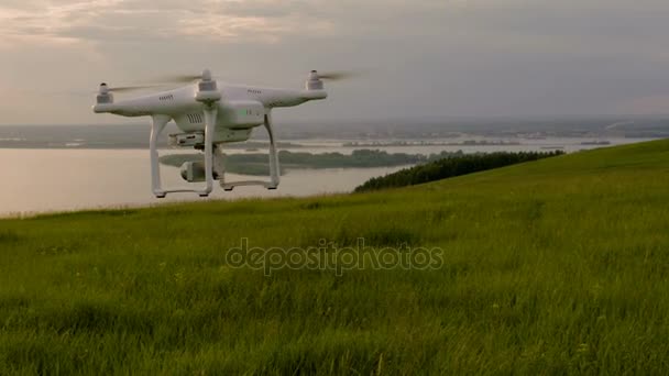 Білий безпілотник літає над пагорбом з зеленою травою і виводиться на відстань на заході сонця — стокове відео