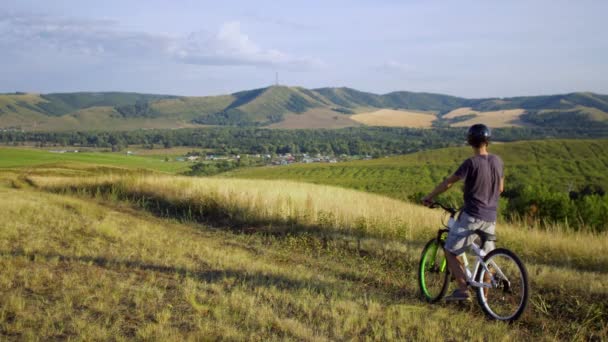Молодой человек на велосипеде едет вдоль полей на закате, останавливается, оглядывается и едет — стоковое видео