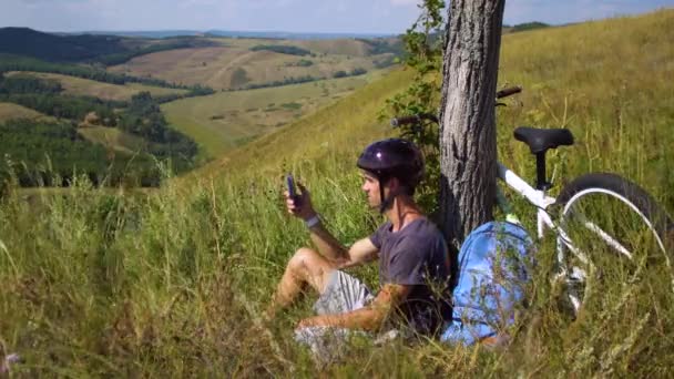 Egy fiatal férfi kerékpár utazás alatt pihen egy fa alatt. Egy mozgatható telefon van a kezében — Stock videók