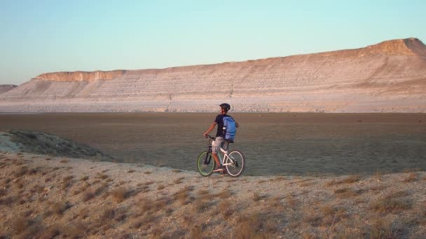 A young man rides a bicycle on a background of a landscape with mountains, stops, drinks water and rides on — Stock Video