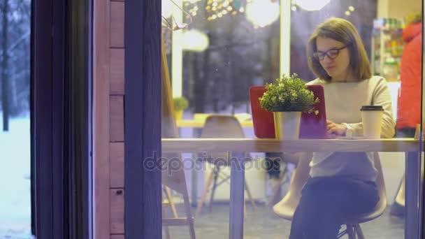Concepto pensativo. Chica joven en un suéter blanco y con gafas, trabajando en un portátil rojo en un café. Vista por la ventana . — Vídeos de Stock