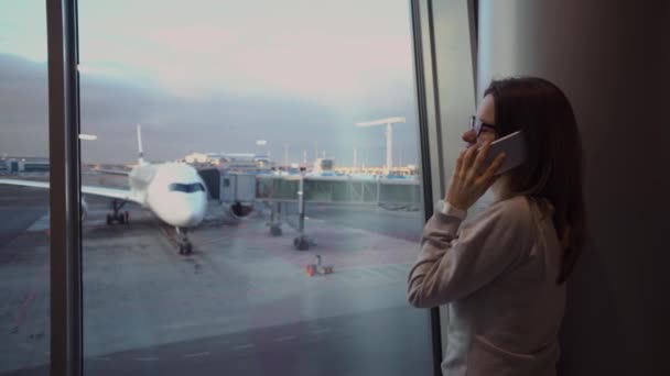 Молодая красивая девушка разговаривает по телефону в терминале аэропорта на фоне самолета — стоковое видео