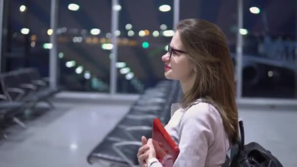 Una chica joven, guapa y sonriente camina por la terminal vacía del aeropuerto, sosteniendo un ordenador portátil y un maletín. — Vídeos de Stock