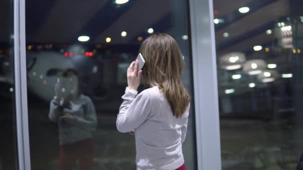 Jong, mooi meisje praten aan de telefoon in een lege luchthaven terminal tegen de achtergrond van een vliegtuig — Stockvideo