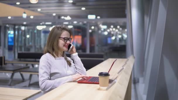 Menina jovem e bonita falando ao telefone em pé em uma mesa com um laptop e uma xícara de café no aeroporto — Vídeo de Stock