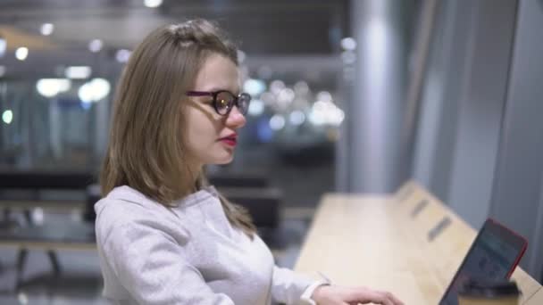 Giovane, bella ragazza che beve caffè da una tazza di carta in piedi a un tavolo con un computer portatile in aeroporto — Video Stock