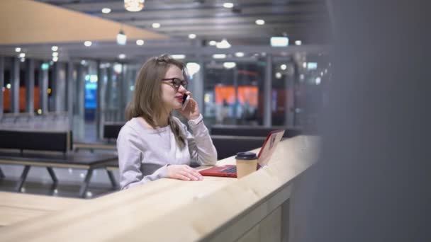 Молодая, красивая девушка разговаривает по телефону, стоя за столом с ноутбуком и чашкой кофе в аэропорту — стоковое видео