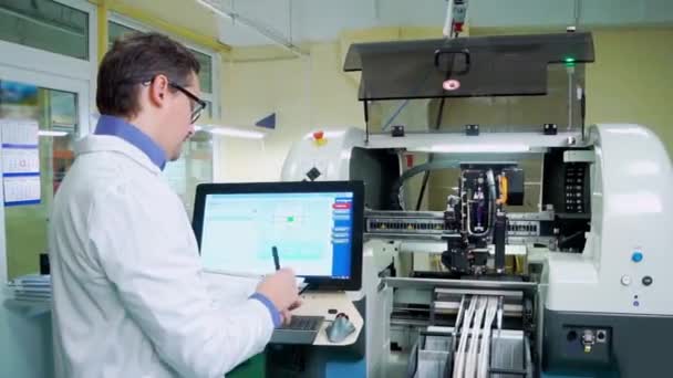 Az Engineer egy fehér köntös és szemüveg dolgozik a Surface Mount Technology gép. PCB feldolgozás CNC megmunkálógépen, elektronikai alkatrészek gyártása High-Tech gyárban