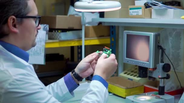 Um homem com um roupão branco e óculos verifica a placa elétrica por defeitos. Departamento de controle de qualidade na produção. fabricação em uma fábrica de alta tecnologia — Vídeo de Stock