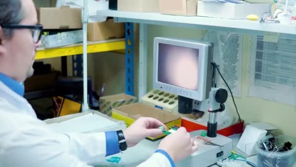 Mężczyzna w białym szlafroku i okularach sprawdza czy na tablicy rozdzielczej nie ma wad przez urządzenie powiększające z wyświetlaczem. Dział kontroli jakości w produkcji. produkcja w fabryce zaawansowanych technologii — Wideo stockowe