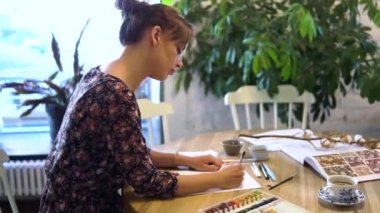 Genç bir kız kafedeki bir masada oturan bir fırçayla kağıda eskizler çiziyor. Moda tasarımcısı, terzisi, animatörü, sanatçısı ya da illüstratörü bir kafede çalışır.