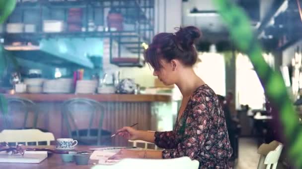 A young girl draws sketches on paper with a brush sitting at a table in a cafe. A fashion designer or tailor, animator, artist or Illustrator works in a cafe — Stock Video