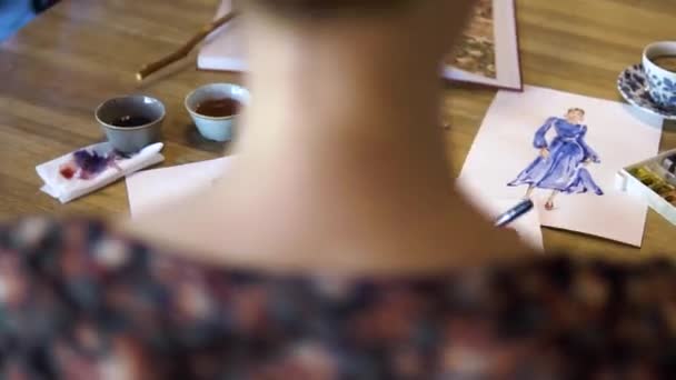 Een jong meisje tekent schetsen op papier met een borstel aan een tafel in een café. Een modeontwerper of kleermaker, animator, kunstenaar of Illustrator werkt in een café — Stockvideo
