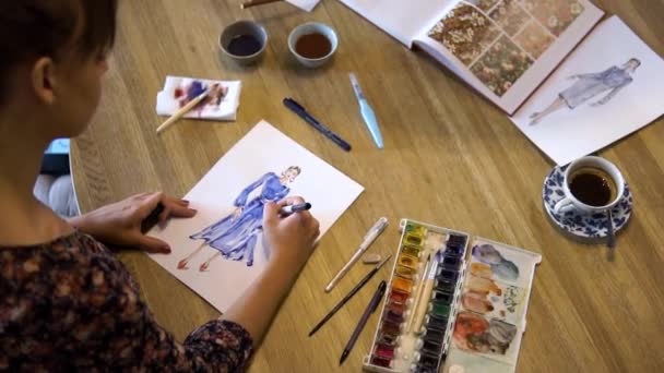Una joven dibuja bocetos en papel con un pincel sentado en una mesa en un café. Un diseñador de moda o sastre, animador, artista o ilustrador trabaja en un café. Vista superior — Vídeo de stock