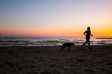 Güzel genç bir kadın gün batımında köpeğiyle plajda koşuyor.