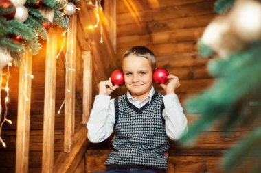 7 yaşındaki beyaz gömlekli ve siyah beyaz örgü yelekli çocuk tahta merdivenlerde oturur ve tatil, kış, aralık aylarında kırmızı Noel toplarıyla oynar. Mutlu ve komik