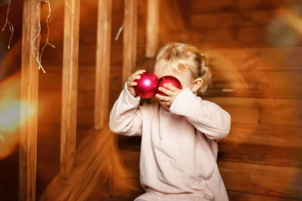 Fille de 3 ans en robe chaude rose s'assoit sur des escaliers en bois et joue avec des boules de Noël rouges pendant les vacances, hiver, décembre. Enfant heureux et drôle dans les lumières de Noël — Photo