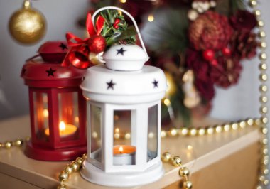 Kırmızı ve Beyaz Yılbaşı Fenerleri ve yanan mumlar. Altın boncuklar ve toplarla süslü Noel süsleri. Arka planda Noel ağacı.