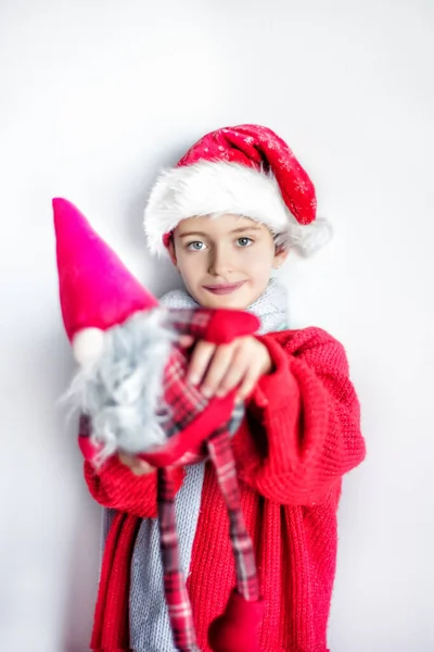 7 ans vieux sourire souriant dans le chapeau de Père Noël, pull rouge, écharpe grise avec dwurf dans le chapeau rouge sur fond neutre. Lumière naturelle. Espace pour le texte — Photo