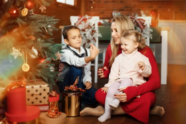 Család - anya, fia és lánya - játszik és mosolyog a padlón közel karácsonyfa ajándékdobozok körül. Nő, fiú és lány - testvér és testvér - boldogok és meglepettek karácsonykor — Stock Fotó