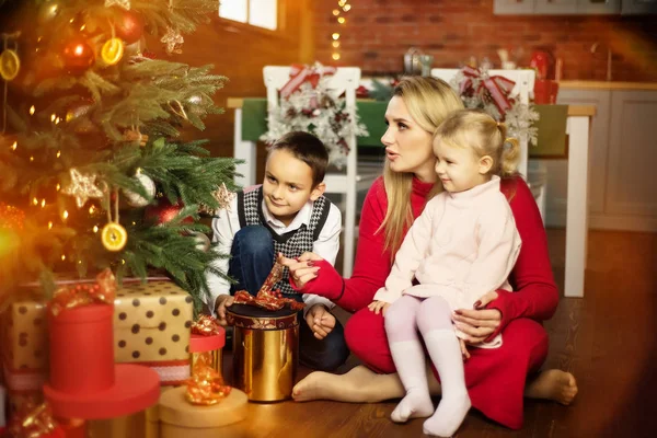 Család - anya, fia és lánya - játszik és mosolyog a padlón közel karácsonyfa ajándékdobozok körül. Nő, fiú és lány - testvér és testvér - boldogok és meglepettek karácsonykor — Stock Fotó