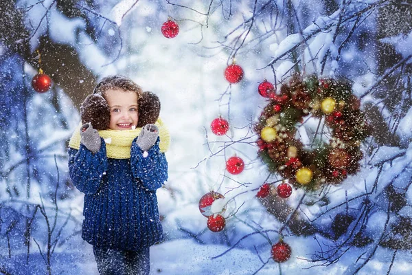 Petite fille souriante dans un pull bleu, écouteurs marron moelleux et une écharpe jaune claque des mains dans une forêt enneigée d'hiver avec des décorations de Noël boules de Noël rouges et couronne sur les branches — Photo