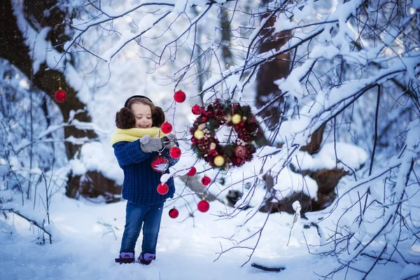 Une petite fille souriante dans un pull bleu, des écouteurs marron moelleux et une écharpe jaune décorent les branches dans une forêt enneigée d'hiver avec des décorations de Noël - boules rouges et couronne de Noël. Vacances — Photo