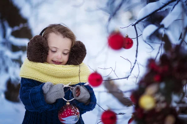身着蓝色套头衫、蓬松的棕色耳机和一条黄色围巾的小女孩微笑着，在冬雪覆盖的森林里装饰着树枝，装饰着圣诞装饰品- -红色的球和圣诞花环。 假日 — 图库照片