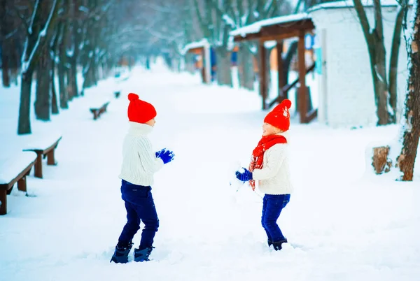 Ragazzina e ragazzo divertente con cappello e sciarpa in maglia rossa e pullover bianco che giocano all'aperto in inverno. I bambini giocano all'aperto in inverno. I bambini si divertono nel periodo natalizio. Fratello e sorella . — Foto Stock