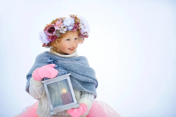 Very sweet beautiful little smiling girl child in pink skirt and mittens, white pullover, gray scarf and floral wreath with old fashioned lantern in hands. Snow is around. Space for text — Stock Photo, Image