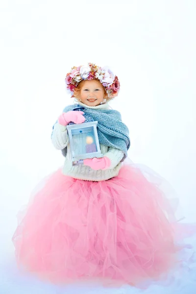 Muy dulce hermosa niña sonriente en falda rosa y manoplas, jersey blanco, bufanda gris y corona floral con linterna antigua en las manos. La nieve está cerca. Espacio para texto — Foto de Stock