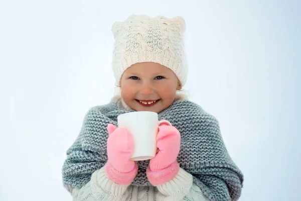 Très douce belle petite fille souriante enfant en jupe rose et mitaines, pull blanc, écharpe grise et chapeau blanc avec tasse blanche dans les mains. Espace pour le texte — Photo