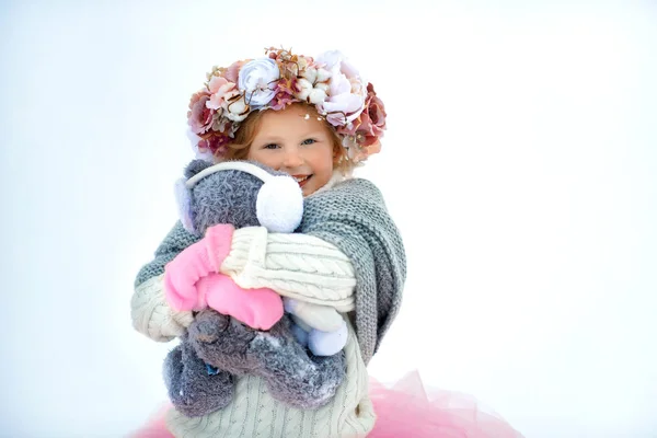 Niña muy dulce hermosa y sonriente en falda rosa y manoplas, jersey blanco, bufanda gris y corona floral con viejo oso taddy en las manos. Nieve alrededor. Espacio para texto — Foto de Stock