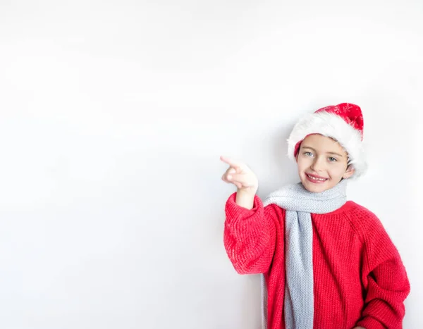Enfant souriant de 7 ans en chapeau de Père Noël, pull rouge et écharpe grise avec un doigt sur la partie gauche de l'image avec de l'espace pour le texte. Un, isolé, affichage — Photo