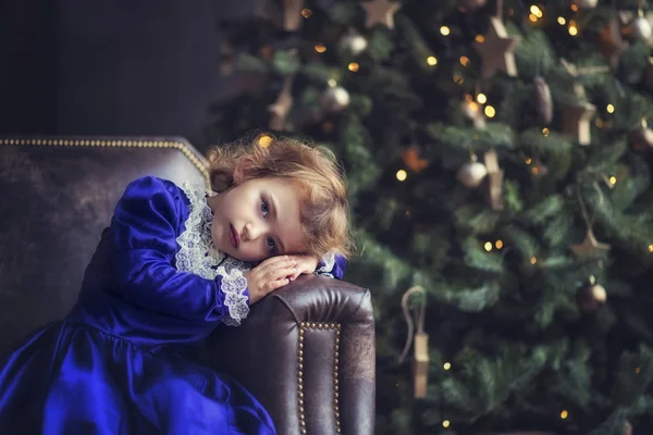 Petite fille de 5 ans en robe violette vintage assise sur un canapé en cuir près de l'arbre de Noël dans la chambre. tête sur les mains. Une fille qui attend le Père Noël et des cadeaux. Noël et Nouvel An veilles — Photo