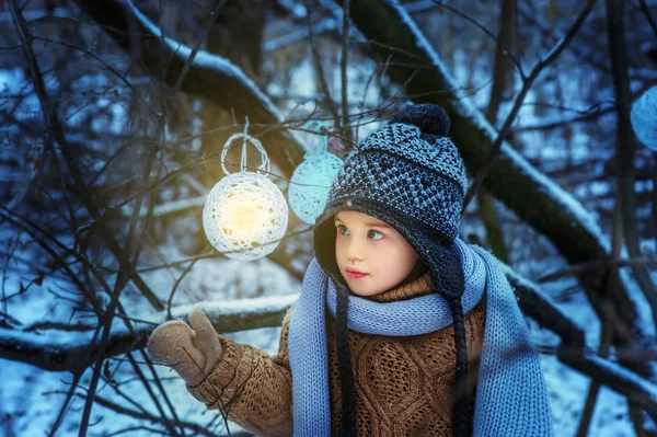 Petite fille de 4 ans dans la forêt enneigée d'hiver regarde boule lumineuse. Enfant en pull marron, écharpe grise et chapeau chaud. Temps de Noël — Photo